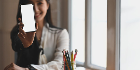 Beautiful woman holding and showing a white blank screen smartphone in her hand while sitting at the working desk over comfortable living room windows as background.