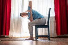 Young Man Practicing Yoga Using Chair, Doing Revolved Chair Pose