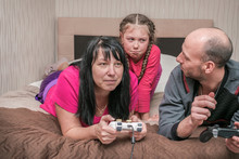 While The Parents Are Playing Video Games, The Child Is Crying. Dad And Mom Do Not Give The Child A Gamepad For A Computer Game. The Family Is Lying On The Bed While Being Quarantined.