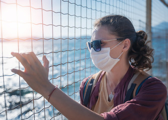 One young girl a woman in a medical mask looks at the sea through an iron grate. Travel ban to the sea, closed border concept