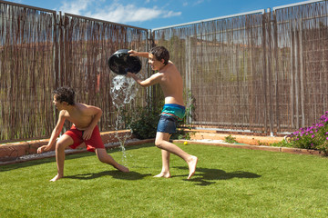 Two boys having a water battle on a hot sunny day in the back yard with a black bucket.