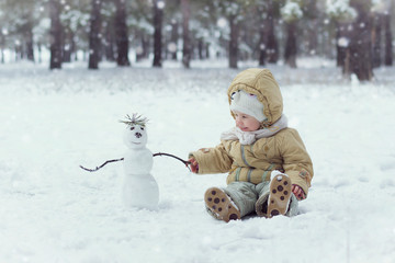 Cute little child sits on snow in winter next to a snowman on a winter day in the forest.