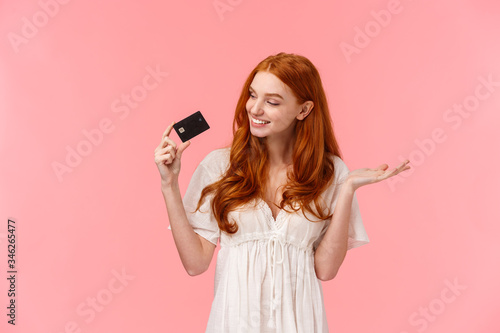 Satisfied cute redhead woman praise her credit card, looking at it pleased and happy, smiling, wasting money on bank account, receive payment and going shopping, standing pink background