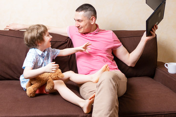 Funny young father sit on computer laptop fool around with cute kid son have fun together, smiling dad and little boy child enjoy stay at home rest on couch with gadgets. Online education and work