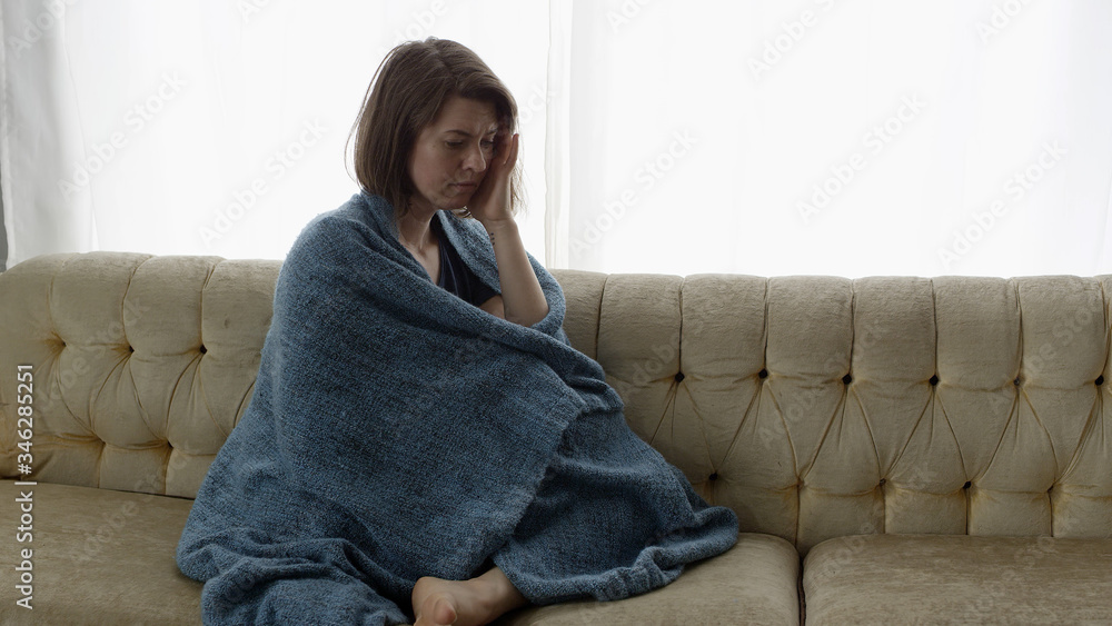 Sad woman sitting on couch wrapped in blanket with hand on her head as ...