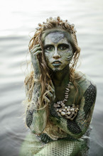 Girl Mermaid Amphibian With Green Skin With Body Art With A Necklace And A Seashell Swimsuit Around Her Neck Holding Her Hand To Her Serious Face In The River Summer In The Water In The Lake