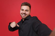 © ViDi Studio - Close up of smiling young bearded guy 20s in black shirt isolated on red background. People lifestyle concept. Mock up copy space. Doing selfie shot on mobile phone, pointing index finger on camera.