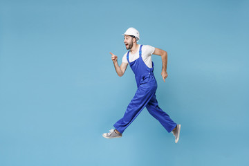 Poster - Side view of excited man in coveralls protective helmet hardhat isolated on blue background. Instruments accessories renovation apartment room. Repair home concept. Jumping, point index finger aside.