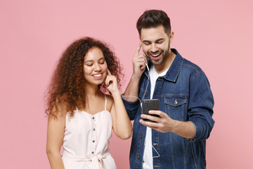 Wall Mural - Funny young couple two friends european guy african american girl in casual clothes posing isolated on pastel pink background. People lifestyle concept. Listen music with earphones using mobile phone.