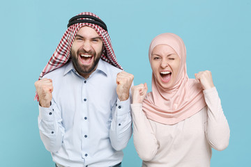 Canvas Print - Joyful young couple friends arabian muslim man wonam in keffiyeh kafiya ring igal agal hijab clothes isolated on blue background in studio. People religious lifestyle concept. Doing winner gesture.