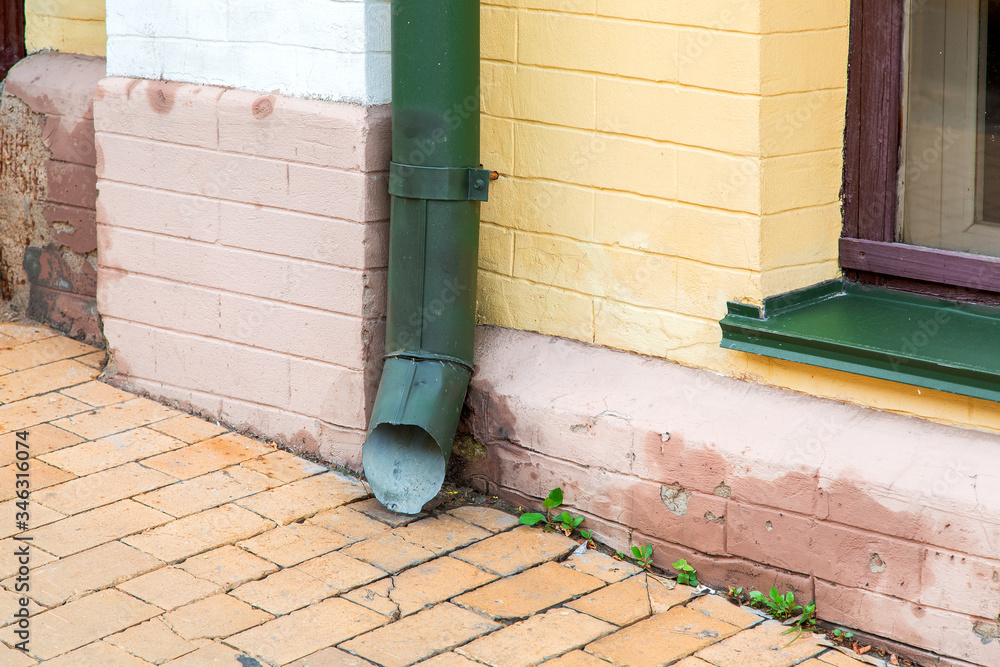 green rain pipe rain system on the facade of the brick house near the stone sidewalk, close up of an architectural element.