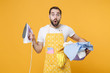 © ViDi Studio - Shocked young man househusband in apron hold basket with clean clothes, iron doing housework isolated on yellow background studio portrait. Housekeeping concept. Mock up copy space. Looking camera.