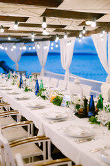 Wall Mural - Wedding dinner table reception. A long table, against the sea, with a white tablecloth, a garland with incandescent light bulbs over it. Wooden white chairs. Flower composition in the center of table