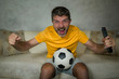 © TheVisualsYouNeed - young excited and crazy happy football supporter man watching soccer game on television at living room sofa holding TV remote celebrating team scoring goal