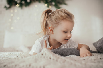 beautiful preschool girl with white wavy shaggy hair tied up in a hairstyle lies on a light cozy bed against a white brick wall, looks at the side and smiles