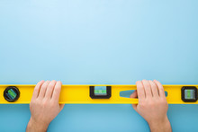 Man Hands Holding Yellow Spirit Level On Light Blue Table Background. Building Tool For Floor, Wall, Ceiling Or Other Surfaces Level Precision. Point Of View Shot. Empty Place For Text. Top Down View.