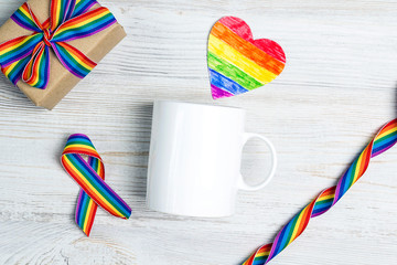 Wall Mural - White coffee mug and gift with rainbow LGBT ribbon on a light wooden background.