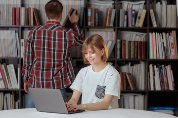 Cute boy and girl students working on a project in the city library, looking for books and typing notes on a laptop.