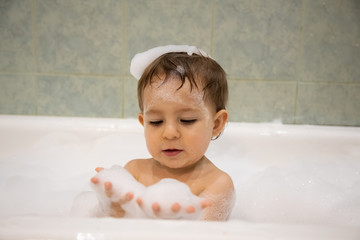 Wall Mural - Cute baby boy taking a bath looking at soap foam in his hands. close up, soft focus, bacground is bathroom in blur