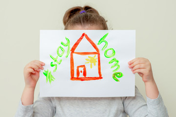 Wall Mural - Child's hands holding a picture of red house and words Stay Home covering her face. Children in quarantine concept.