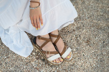 Female Feet In Sandals On Sandy Greek Shore. Girl Resting On Beach