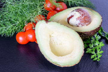 Wall Mural - Halves of fresh big avicado with tomatoes cherry and heap of dill and parsley on the black background. Ketogenic diet food avocado.