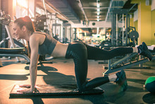 Fit Athletic Woman Exercising Down On The Floor Balancing On Her Arms And One Knee With The Other Leg Raised Up Behind Her In The Air At Gym. Side View.