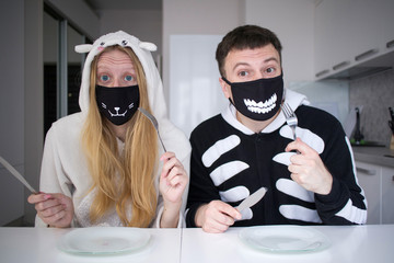 Wall Mural - young brunette guy in black with white pajamas with a hood (kigurumi) and a blonde girl in white pajamas (kigurumi) in unusual black masks with a white print have fun on self-isolation in the kitchen