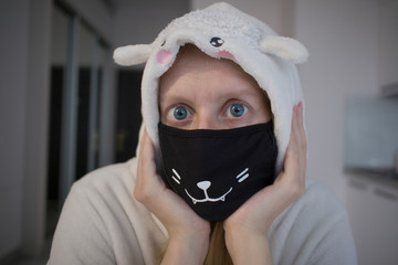 Wall Mural - blonde girl in white pajamas (kigurumi) in an unusual black mask with a white print has fun on self-isolation in the kitchen