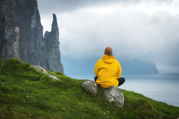 Wall Mural - Tourist in yellow jacket looks at Witches Finger cliffs from Trollkonufingur viewpoint. Vagar island, Faroe Islands, Denmark.