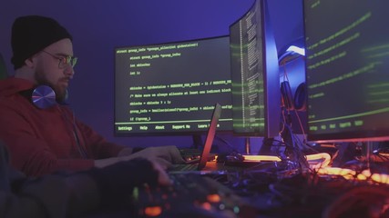 Lockdown of bearded Caucasian man wearing glasses and hat and his unrecognizable colleague sitting together at multiple computers and working