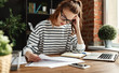 © JenkoAtaman - Young dedicated woman revising papers while working on project at laptop in loft office.