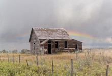 Old Dilapidated Farm House Behind A Barbed Wire Fence Under A Grey Sky With A Rainbow