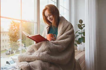 pretty thoughtful smiling young girl reading book and drinking morning coffee at home sitting next to the window wrapped in warm comfy blanket