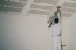 Man dressed in white, a painter, is applying putty or stucco to a dry wall installation in a house. Smoothing surfaces of a drywall application, reaching high up to the roof.
