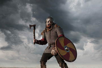 Warrior Viking in full arms with axe helmet and shield on dark sky background. Medieval scandinavian berserk red beard and fur costume attacks enemy. Concept historical photo