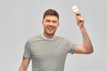 Wall Mural - eco living, inspiration and sustainability concept - smiling young man in striped t-shirt holding lighting bulb over grey background