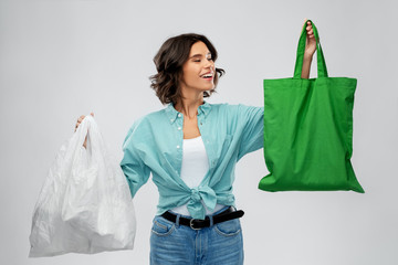 consumerism, sustainability and eco living concept - portrait of happy smiling young woman in turquoise shirt and jeans with plastic and green reusable canvas bag for food shopping on grey background