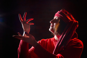 Wall Mural - Portrait of middle age woman in red sweater and the turban of wizard or sorcerer on black background. Actress in a magical, fantastic, fairy-tale movie. Female model posing at studio with low light