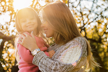 Portrait of young tender mother hugging her little cheerful daughter while standing outdoors. Beautiful woman and little girl embracing and having fun together in autumn park. Parenthood love, family