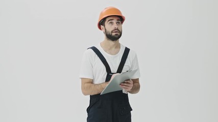 Sticker - Concentrated young bearded man builder isolated over white wall background in helmet writing notes in clipboard.