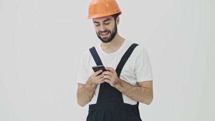 Sticker - Cheery optimistic young bearded man builder isolated over white wall background in helmet using mobile phone