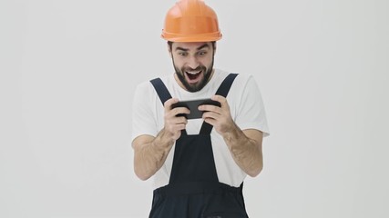 Sticker - Excited emotional young bearded man builder isolated over white wall background in helmet play games by mobile phone