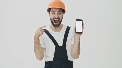 Sticker - Positive cheery young bearded man builder isolated over white wall background in helmet showing display of mobile phone