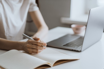 Young woman works on a laptop and takes notes in a notebook. Work in home office.