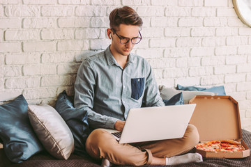 Busy businessman freelancer using laptop for distance work from home office, eat tasty pizza. Stylish hipster man student in glasses surf internet on computer, study remotely. Cozy workplace on couch