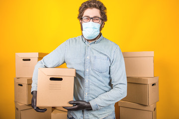 Wall Mural - Bearded man courier in a medical mask holding a corton box with a parcel in his hand. Standing among many corton boxes on a yellow background