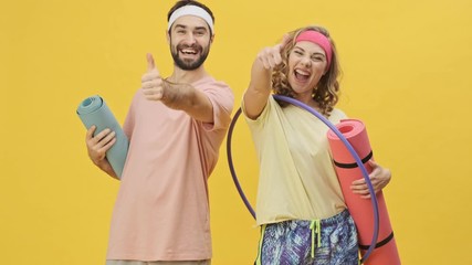 Poster - A happy attractive young athletic couple man and woman in sportswear are showing thumb-up gesture while holding sports equipment isolated over a yellow background in studio