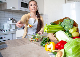 Wall Mural - Young brunette woman making healthy food on the dinner table in the kitchen. Female cooking fresh vegan salad with green vegatable