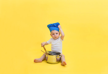 A little boy looks at the camera and a chef's hat with a pot and a wooden spatula spoon. A boy in a white bodysuit pulls on a cap